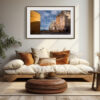 A cosy living room with a low wooden sofa, brown and white cushions, a round ottoman, a potted plant, and a large window. The 'Bastia Old Town' Print featuring a Mediterranean street scene hangs on the wall.