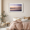 A cosy bedroom features a beige duvet, neutral cushions, a leafy plant, and the 'False Bay Sunset' Print framed on the light wall above the bed.