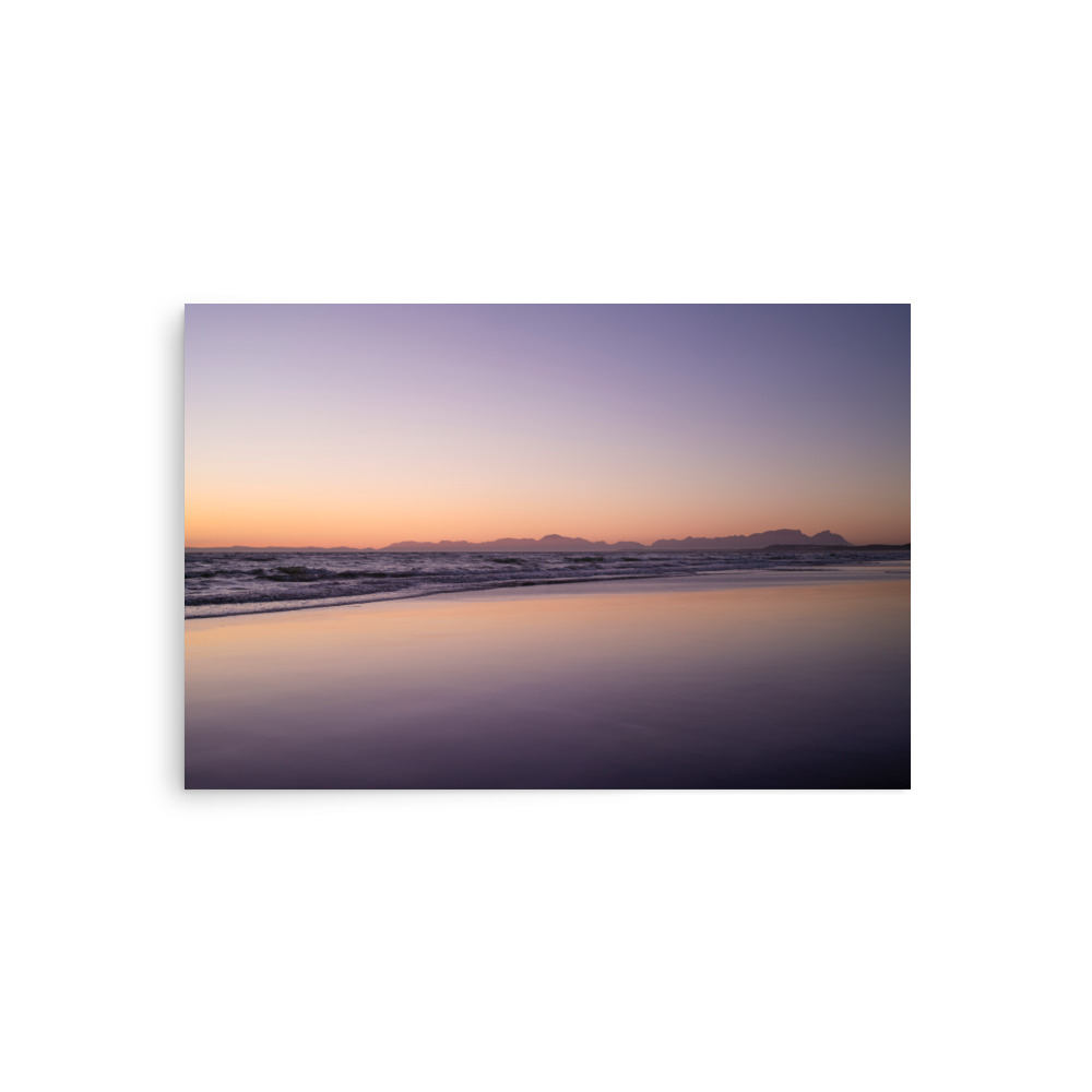 Experience the tranquil beauty of the 'False Bay Sunset' Print, where gentle ocean waves meet sandy shores beneath pastel purple, orange, and yellow skies at sunset, with distant mountains gracing the horizon.