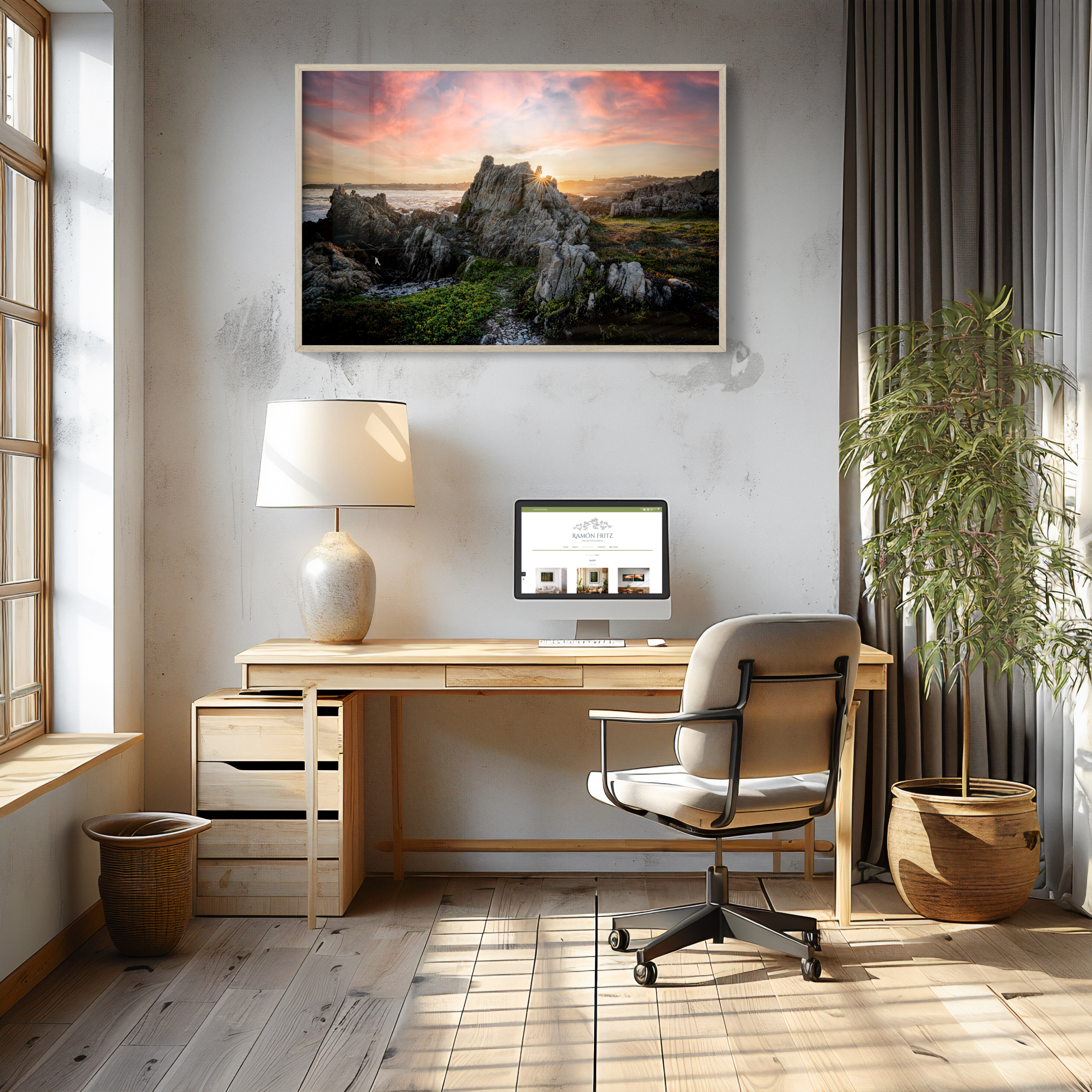A modern home office with a wooden desk, chair, computer, lamp, potted plant, and the 'Van Dyk's Bay' (H) Print—showing rocky cliffs at sunset—framed above the desk. Sunlight streams in through large windows.