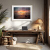 A cosy home office features a wooden desk, chair, laptop, basket, a large framed 'Island Horizon' Print of a sunset beach, tall leafy plant, woven lamp, and natural sunlight shining through an arched window.