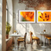 A cosy café with white tables, wooden chairs, a potted plant near a sunlit window, and two vibrant 'Lilium' Diptych Prints displayed on a rustic brick and plaster wall.