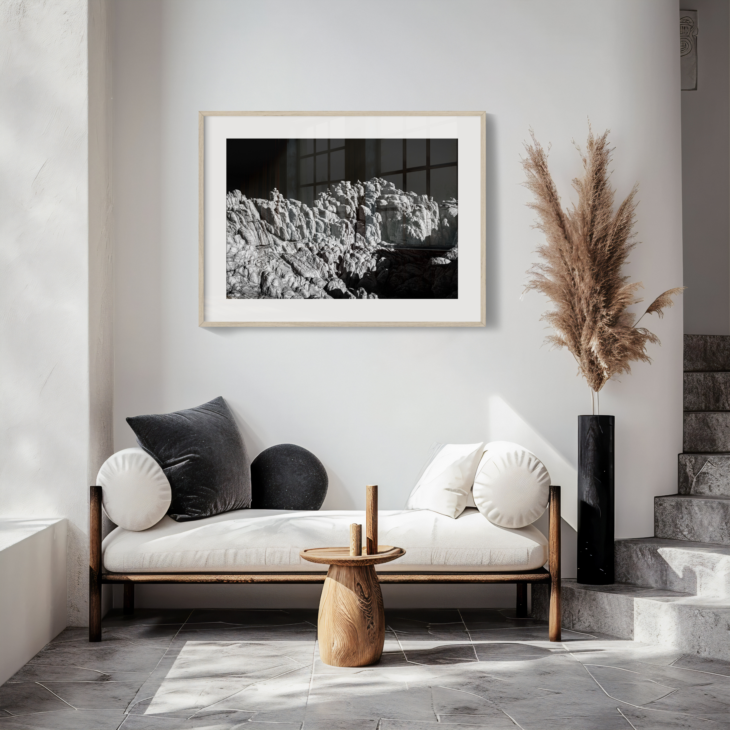 A modern, minimalist living space with a white cushioned bench, round cushions, a wooden table, a tall black vase with pampas grass, and the 'Lunar' (H) Print—a large black-and-white lunar-inspired photograph—on the wall.