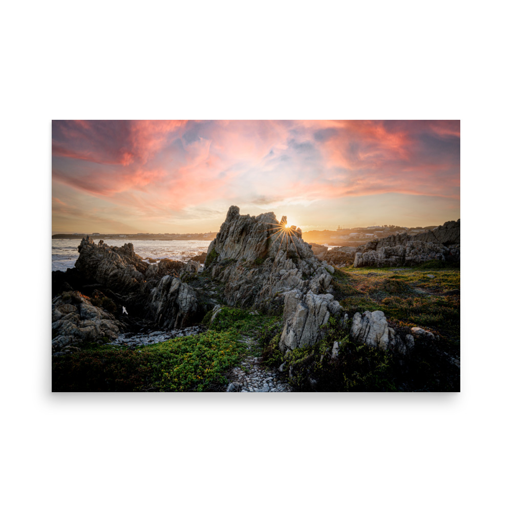 ‘Van Dyk’s Bay’ (H) Print captures a rocky coastal landscape at sunset, where dramatic clouds and sun rays illuminate jagged rocks and lush green vegetation by the sea.