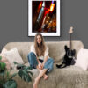 A woman with long hair sits barefoot on a beige sofa beside a black electric guitar. A green plant is to her left, and the 'Nights in Marseille' Print glows on the grey wall behind her above light cushions.