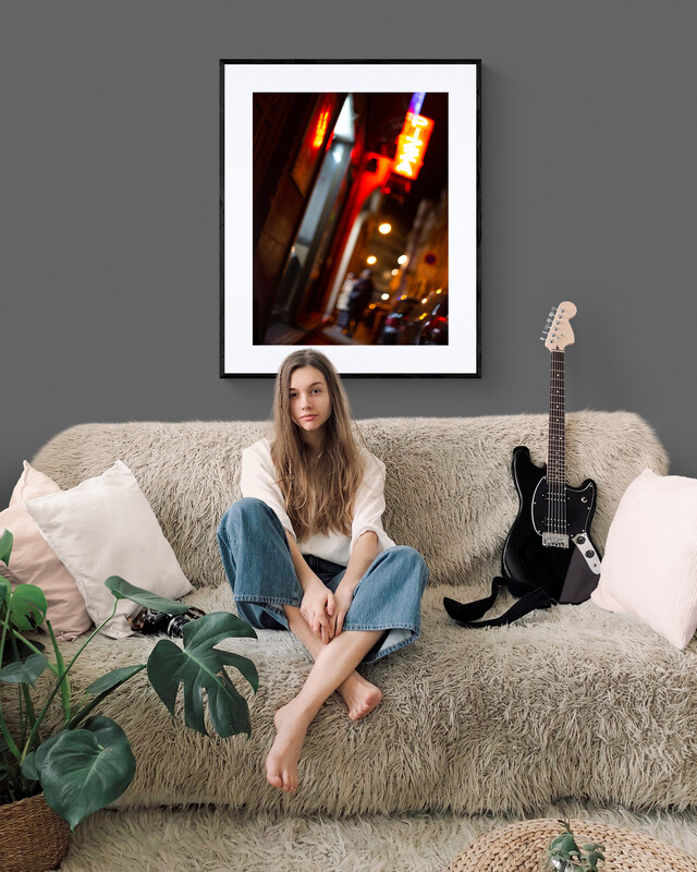 A woman with long hair sits barefoot on a beige sofa beside a black electric guitar. A green plant is to her left, and the 'Nights in Marseille' Print glows on the grey wall behind her above light cushions.