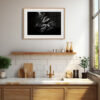 A modern kitchen features a white tiled splashback, gold tap, wooden chopping boards, and plants. Above the sink hangs the 'Vestige' Print—an abstract black and white photo—while sunlight streams through a large window.