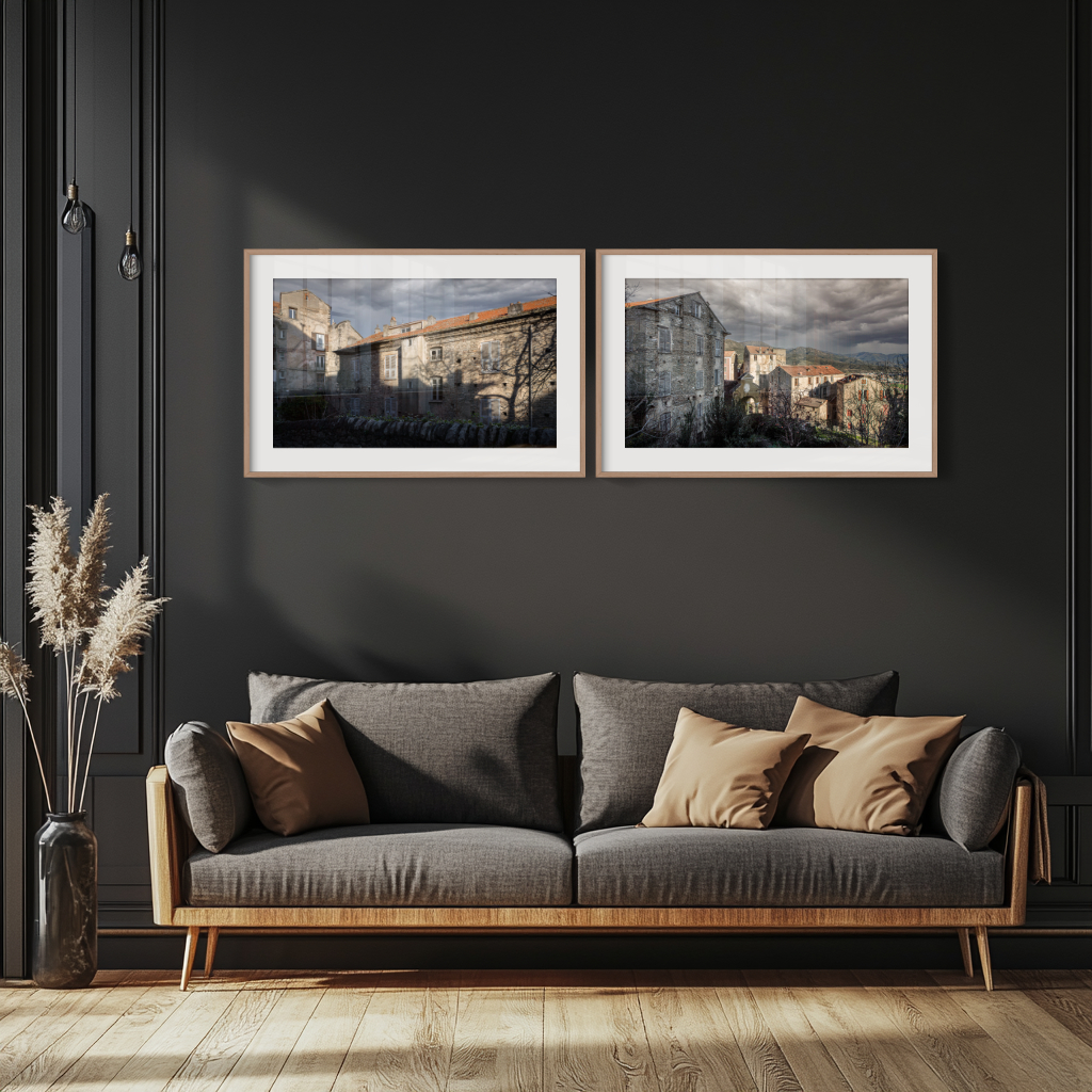 A modern living room features a dark wall, grey sofa with beige and brown cushions, pampas grass in a vase, and the 'Corte' Diptych Prints—two framed landscape photographs of stone buildings in Corte—displayed above the sofa.