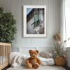 A cosy nursery features a cot, plush bear, soft blankets, plants, and a framed 'Neighbourhood Watch' print showing a cat peering from a green-shuttered window in an old building.