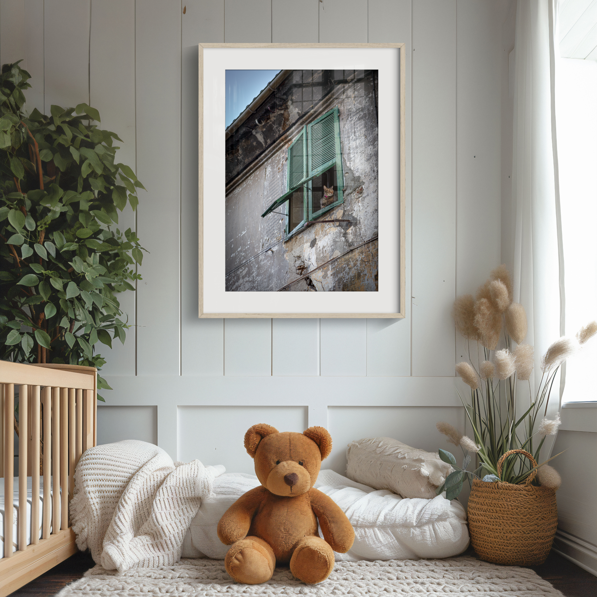 A cosy nursery features a cot, plush bear, soft blankets, plants, and a framed 'Neighbourhood Watch' print showing a cat peering from a green-shuttered window in an old building.