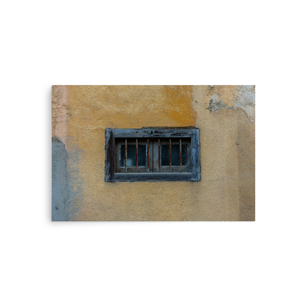 The 'Window of Old Town Bastia' Print features a small rectangular window with black metal bars set in a weathered yellow wall, highlighted by flaking paint and a textured, rustic finish.