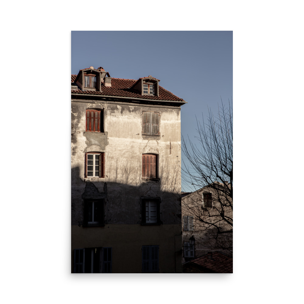 Bring home the charm of the 'Weathered Light' Print, featuring a sunlit flat building with closed wooden shutters and a red-tiled roof beside bare branches under a clear blue sky.