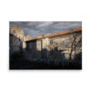 The 'Corte II' Print depicts old stone buildings with closed shutters illuminated by dramatic sunlight, shadows of bare tree branches extending across the walls, and a stone fence in front beneath a cloudy sky.