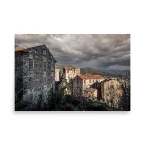 The 'Corte I' Print features old stone buildings with shuttered windows on a hillside beneath a dramatic cloudy sky, distant mountains in the background, and sparse leafless trees and bushes in the foreground.