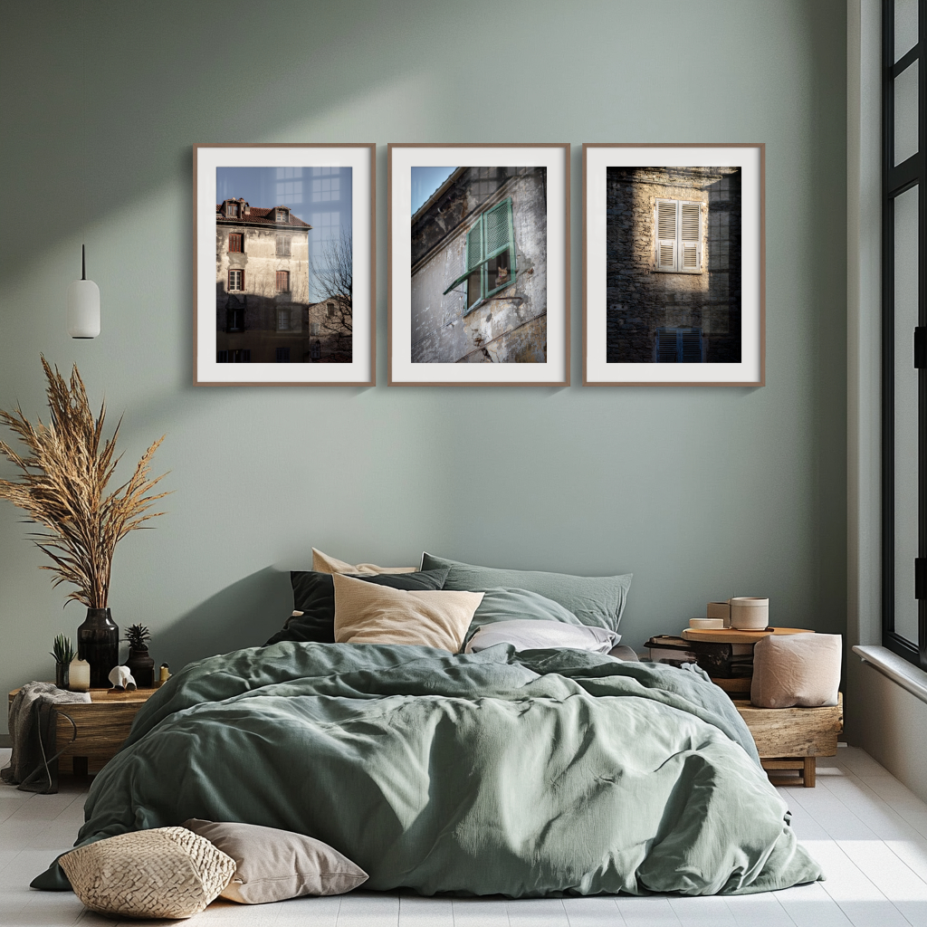 A modern bedroom features the 'Shutters of Corte' Print Collection above the bed, with a green duvet, neutral pillows, dried plants in a vase, and soft green walls. Large windows fill the space with natural light.