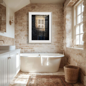 A rustic bathroom with stone walls, a white freestanding bath draped with a towel, and the 'Sunset Shutters' Print framed on the wall. Sunlight pours in, illuminating a wicker basket and the basin with its mirror.