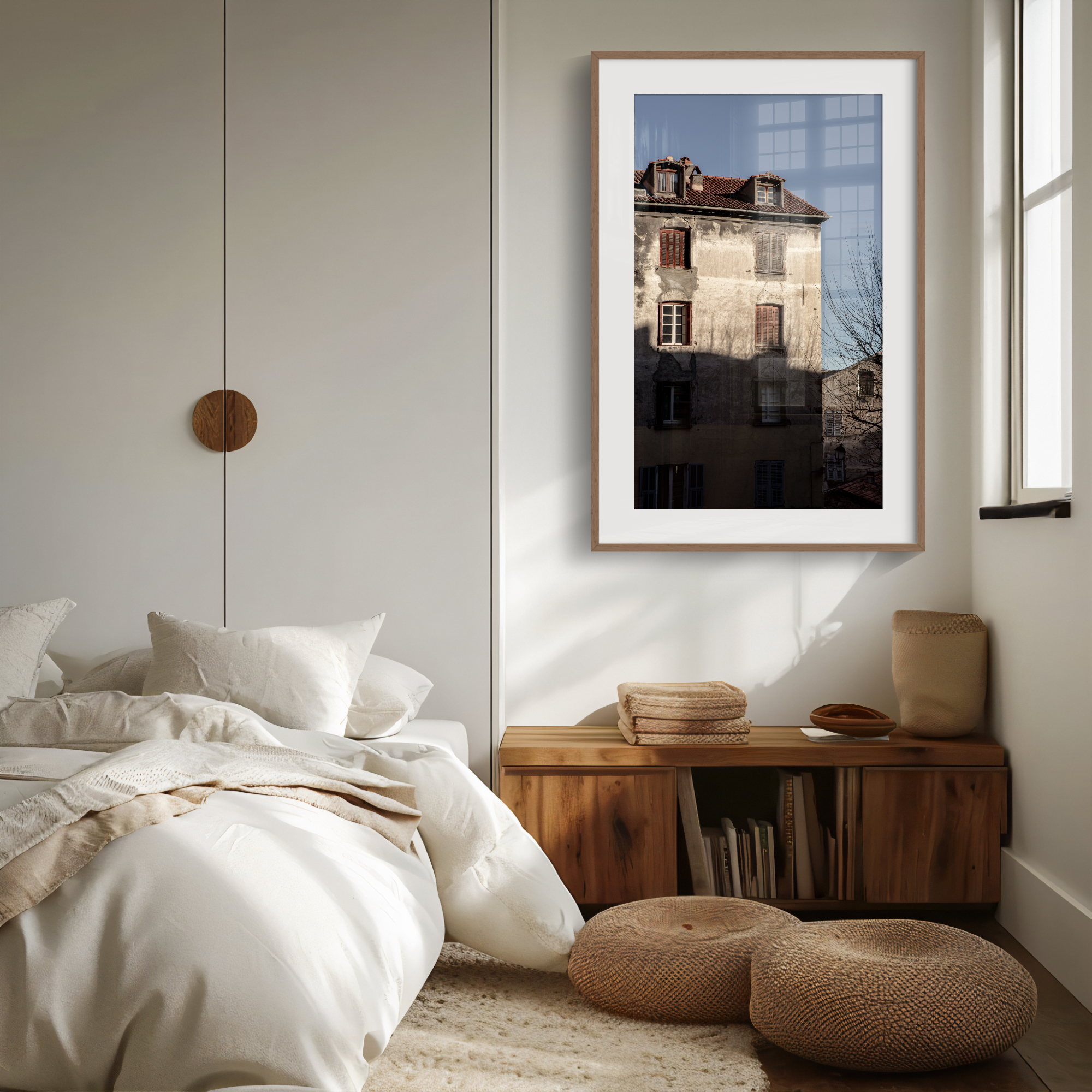A cosy bedroom with neutral tones features an unmade bed, a wooden bench with towels, a round pouffe, and the 'Weathered Light' Print—a large framed photo of a sunlit European building—hanging on the wall near a window.